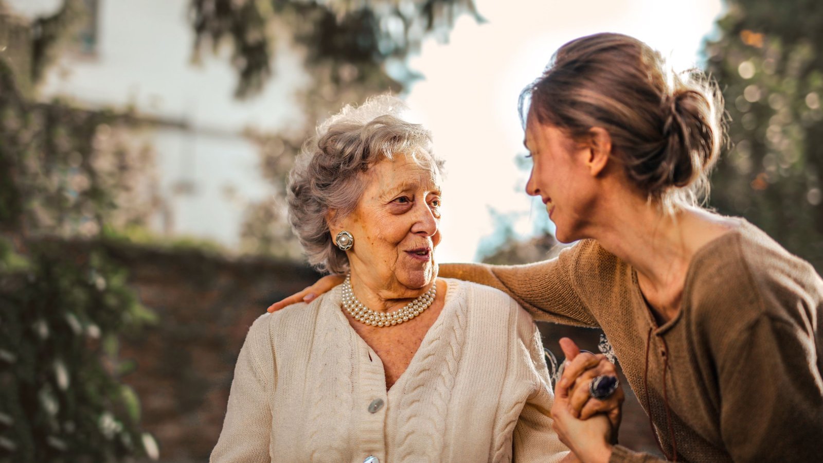 Low angle of cheerful aged lady and adult woman in casual clothes holding hands and smiling at each other while having meeting on yard against blurred green trees and facade of country house in suburb