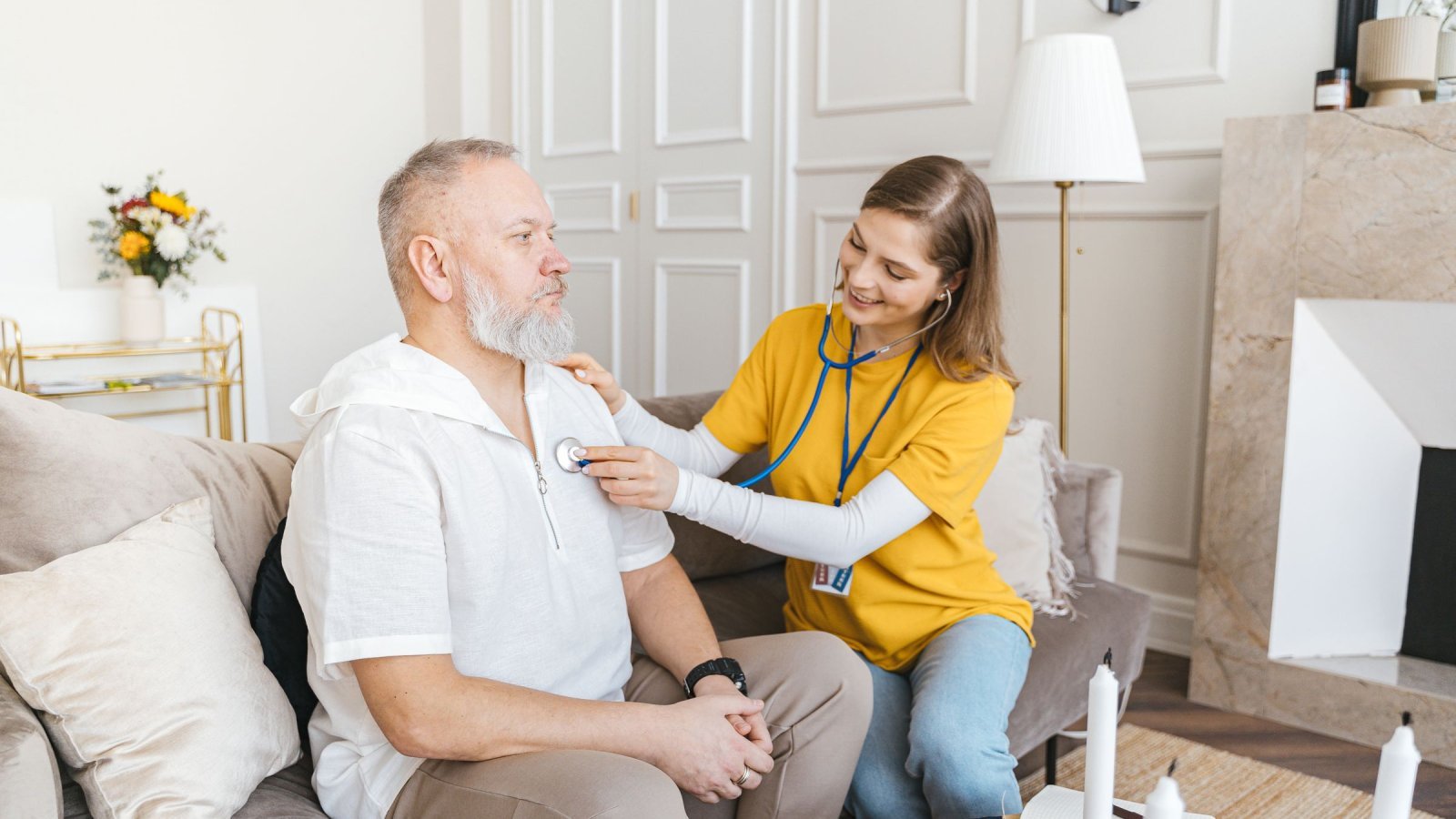 A nurse uses a stethoscope for a home check-up on a senior adult in a cozy living room.