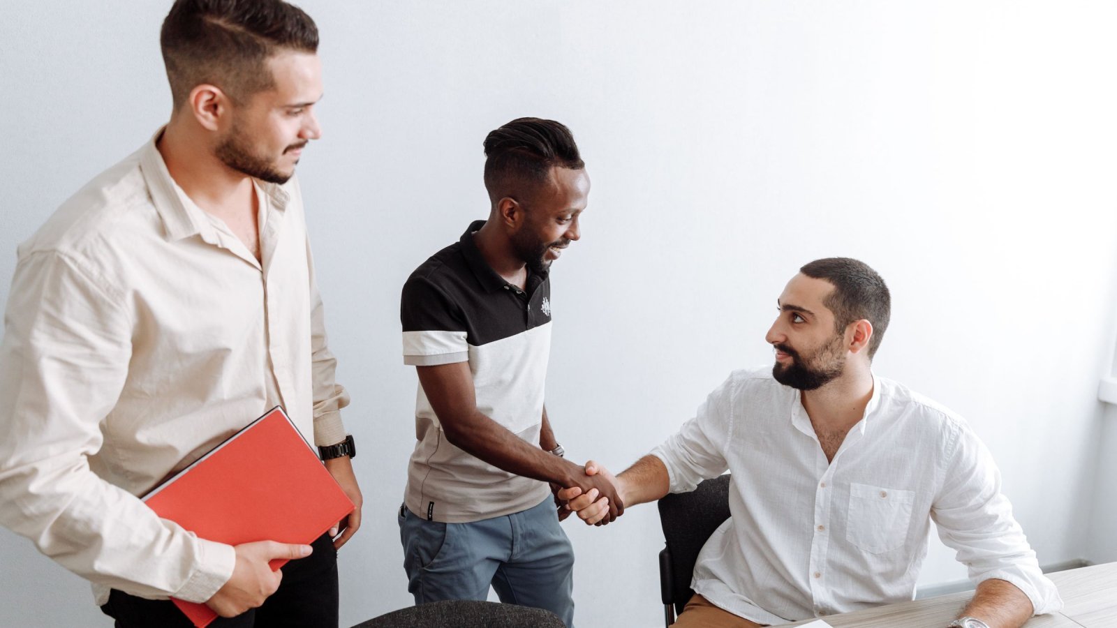 Three men in an office setting, shaking hands, representing teamwork and diversity.